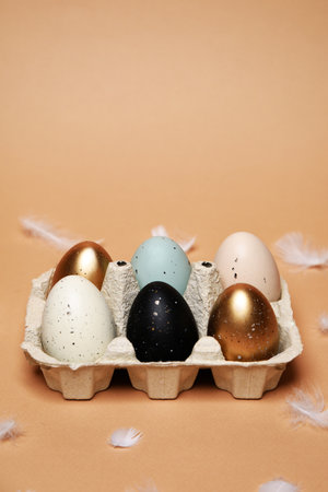 Easter is coming, a tray with painted eggs, on a nude background. Celebrations and traditions. Blue, gold and white with black egg, chicken feathers flying. Plastic eggs, place for text.の写真素材