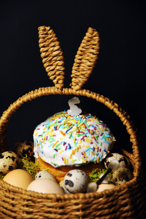 Easter decorations in a basket with rabbit ears. Easter cake and eggs on moss, quail eggs and feathers on a black dark background. A small figure of a rabbit on an Easter cakeの写真素材