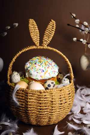 Easter holidays, cute basket with bunny ears on brown background. Easter cakes with colored sprinkles, colored golden eggs, and pussy willow decorations. Nest with eggs, Traditionsの写真素材