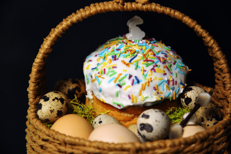 Easter decorations in a basket with rabbit ears. Easter cake and eggs on moss, quail eggs and feathers on a black dark background. A small figure of a rabbit on an Easter cakeの写真素材