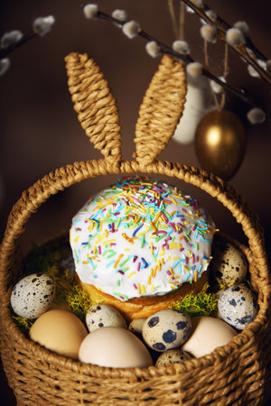 Easter holidays, cute basket with bunny ears on brown background. Easter cakes with colored sprinkles, colored golden eggs, and pussy willow decorations. Nest with eggs, Traditionsの写真素材