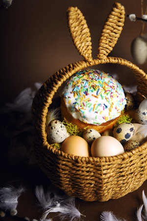 Easter holidays, cute basket with bunny ears on brown background. Easter cakes with colored sprinkles, colored golden eggs, and pussy willow decorations. Nest with eggs, Traditionsの写真素材