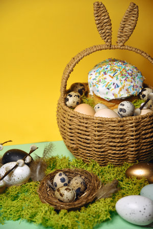 Easter celebration. Easter decorations in a basket with rabbit ears. Easter cake and eggs on moss, quail eggs and feathers on a yellow background. Traditions and religionの写真素材