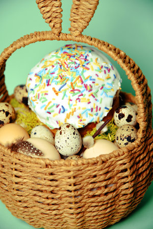 Easter decorations in a basket with rabbit ears. Easter cake and eggs on moss, quail eggs and feathers on mint green backgroundの写真素材