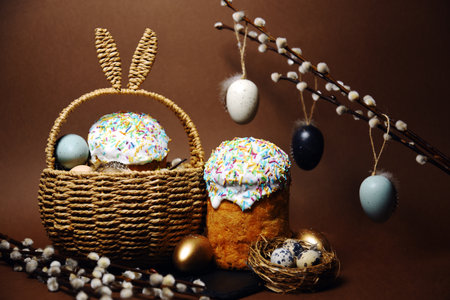 Easter holidays, cute basket with bunny ears on brown background. Easter cakes with colored sprinkles, colored golden eggs, and pussy willow decorations. Nest with eggs, Traditionsの写真素材