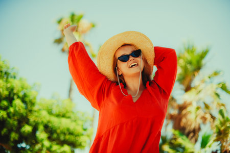 Fashion photo of a girl in black glasses with a chain and a large straw hat, a red dress on a background of palm trees. Wind in the hair of the model, summer photo, vacation, rest. girl enjoy lifeの写真素材