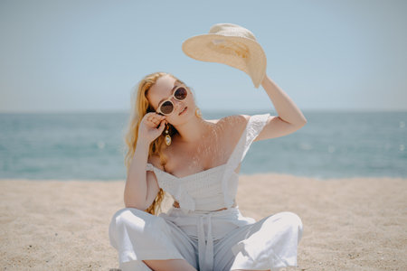Happy girl in white outfit sits on sand against the backdrop of the sea or ocean beach. Woman smiles and laughs, vacation and joy. Fashion model, beautiful jewelry, earrings, hat, sun glasses.の写真素材