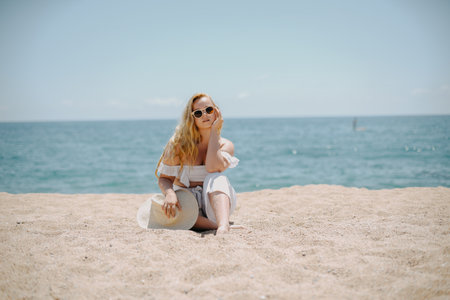 Blonde girl in a hat and sunglasses sits on the sand on the seashoreの写真素材