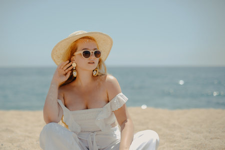 Happy girl in white outfit sits on sand against the backdrop of the sea or ocean beach. Woman smiles and laughs, vacation and joy. Fashion model, beautiful jewelry, earrings, hat, sun glasses.の写真素材