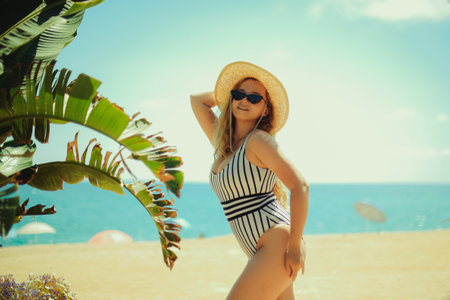 A blonde posing in the summer on the beach, a woman in a hat and fashionable sun glasses stands on the sand. Ocean or sea on the background. Rest under the palm trees.の写真素材