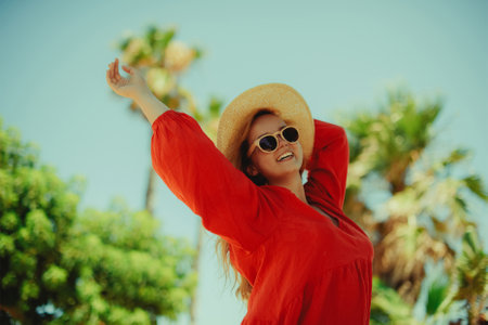Happy girl in a red dress on vacation on the background of palm trees laughing and smiling. vacation. Woman in sun glasses and big hat enjoy life.の写真素材