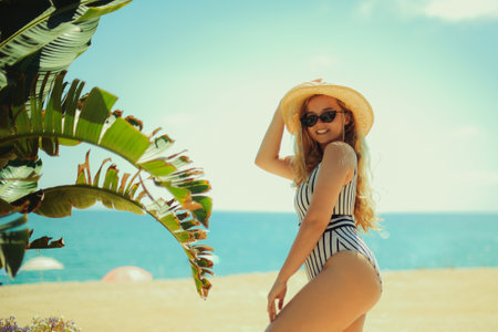 A blonde posing in the summer on the beach, a woman in a hat and fashionable sun glasses stands on the sand. Ocean or sea on the background. Rest under the palm trees.の写真素材