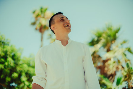 A handsome young man in a white shirt stands against a background of palm trees. Man on vacation, handsome guy summer photo, vacation.の写真素材