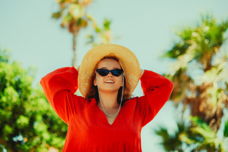 Happy girl in a red dress on vacation on the background of palm trees laughing and smiling. vacation. A woman in sun glasses and a big hat enjoys life and dancesの写真素材