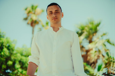 A handsome young man in a white shirt stands against a background of palm trees. Man on vacation, handsome guy summer photo, vacation.の写真素材