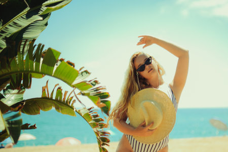 A blonde posing in the summer on the beach, a woman in a hat and fashionable sun glasses stands on the sand. Ocean or sea on the background. Rest under the palm trees.の写真素材