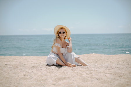 beautiful blonde woman in hat and sunglasses sitting on sand on the beachの写真素材