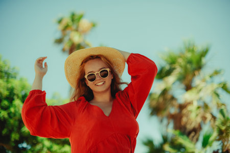 Happy girl in a red dress on vacation on the background of palm trees laughing and smiling. vacation. Woman in sun glasses and big hat enjoy life.の写真素材