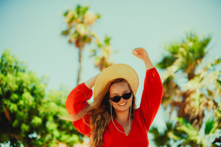 Happy girl in a red dress on vacation on the background of palm trees laughing and smiling. vacation. A woman in sun glasses and a big hat enjoys life and dancesの写真素材