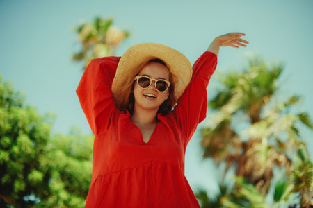 Happy girl in a red dress on vacation on the background of palm trees laughing and smiling. vacation. Woman in sun glasses and big hat enjoy life.の写真素材