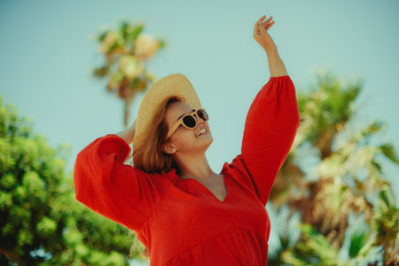 Happy girl in a red dress on vacation on the background of palm trees laughing and smiling. vacation. Woman in sun glasses and big hat enjoy life.の写真素材