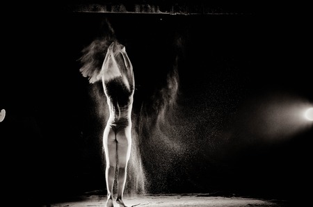 The girl with the flour on the body stretches the arms up with thrown flour on black background black and white image.の写真素材