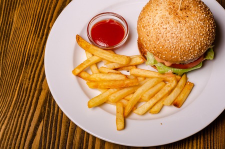 Tasty burger and french fries on plate, on wooden table background. Unhealthy food concept.