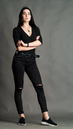 Portrait of young brunette woman sitting on high wooden chair with crossed legs. Fashionable stylish girl wearing wide leg pants. Studio shoot on gray backgroundの写真素材