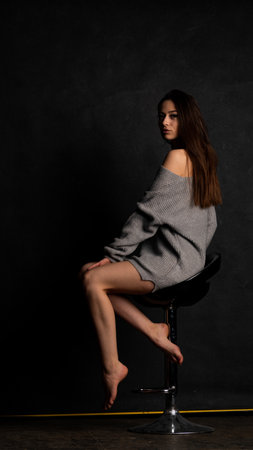 Beautiful young woman in a gray sweater sits on a bar stool on a black backgroundの写真素材