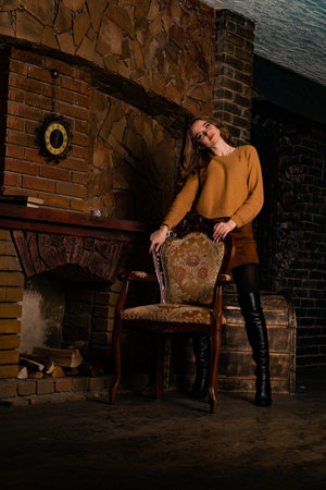 Beautiful young woman in a brown sweater and black boots sits on an armchair near the fireplace.の写真素材