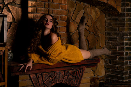Beautiful woman in a yellow sweater sitting on a fireplace in a dark roomの写真素材