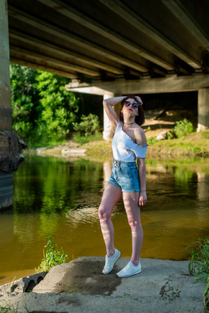 A young brunette woman in a white T-shirt and denim shorts stands by the river under a bridge.の写真素材