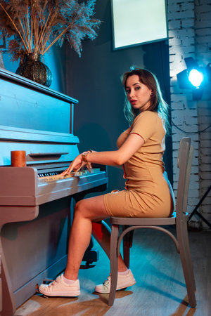 Young beautiful slim brunette girl with long curly hair in long beige dress with open shoulders stands near white grand piano in the roomの写真素材