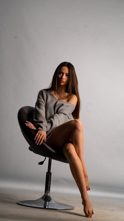 Beautiful girl in a gray sweater sits on a chair on a gray backgroundの写真素材
