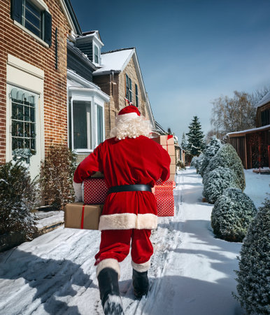 Santa Claus carrying a stack of wrapped presents, walking on a snow-covered path between houses on Christmas Eveの写真素材