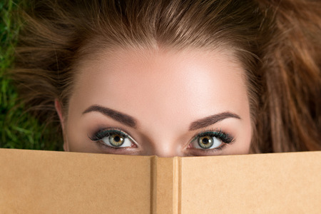 Beautiful young woman looking over book cover laying on grass. Summertime, leisure and education concept Top viewの写真素材