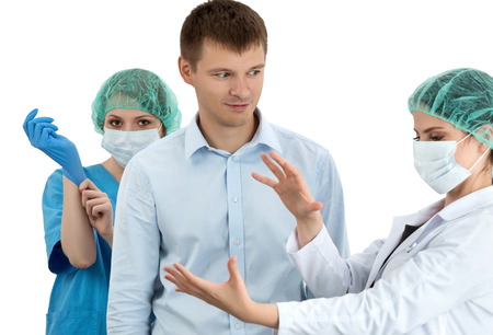 Female doctor in cap and face mask putting on blue protective glove standing behind the scared patient. Her colleague preparing him to the examination. Prostatitis prophylaxis. Men health. Healthcare, medical, surgery and team work conceptの写真素材