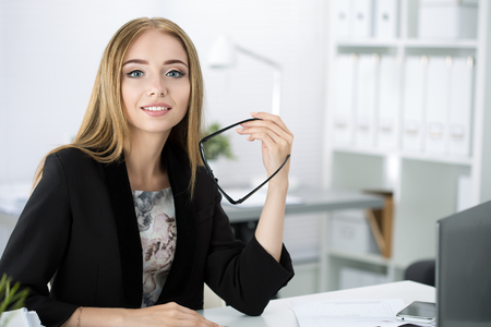 Beautiful pensive businesswoman sitting at her working place at the office and holding glasses in her hand.の写真素材