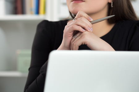 Close up view of woman sitting at laptop, holding pen and thinking of something or listening to someone. Education seminar or blog writing concept.の写真素材