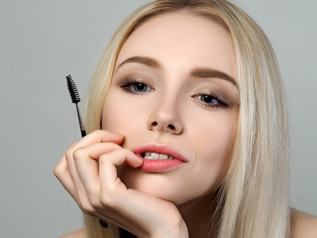 Young beautiful thoughtful blonde woman looking at the mirror and holding a brow brush. Cosmetics, beautician and makeup mistakes conceptの写真素材