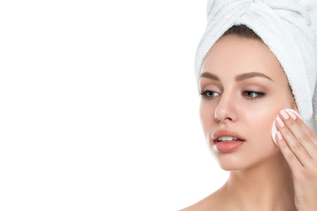 Portrait of young beautiful woman with towel on her hair cleaning makeup from her face with cosmetic pad isolated over white background. Cleaning face, perfect skin, skincare and cosmetology conceptの写真素材