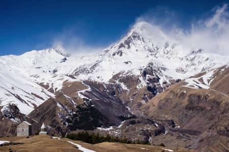 Small orthodox church with Kazbegi mountain covered in snow in the background, clear blue sky