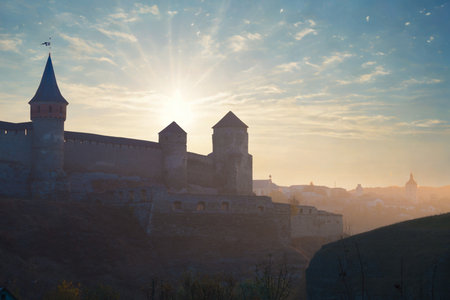 A majestic ancient castle with spires is dramatically silhouetted against a warm, glowing sunrise,...