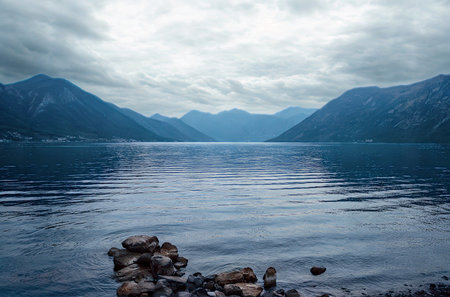 A vast, calm lake reflects a dramatic, cloudy sky between imposing, misty mountains.の写真素材