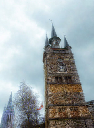 A tall, ancient stone tower with multiple spires, partially obscured by a bare tree, under a dramatic cloudy sky.の写真素材