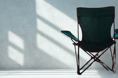 An empty black folding chair sits against a textured wall, illuminated by stark sunlight casting geometric shadows.の写真素材