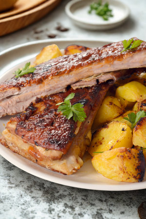 A close-up shot of succulent grilled pork ribs and golden roasted potatoes, garnished with fresh herbs, presented on a white plate.の写真素材