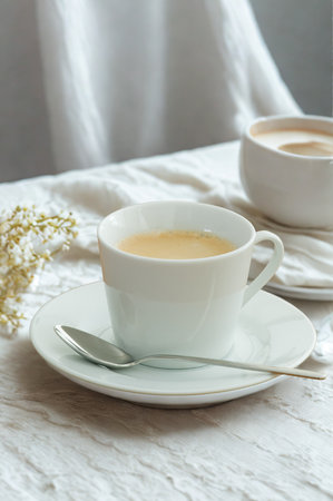 A white teacup filled with a light-colored beverage sits on a matching saucer with a spoon. Soft fabric drapes in the background.の写真素材