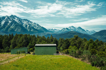 Two contemporary green structures stand in a vibrant green field, surrounded by dense forest and majestic snow-capped mountains under a cloudy sky.の写真素材