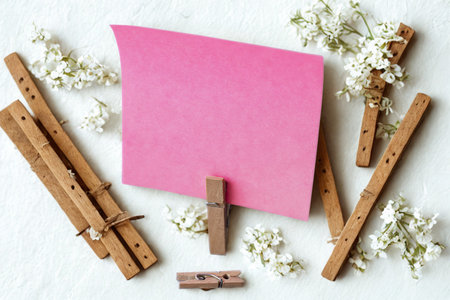 A pink square paper note is held upright by wooden clothespins, with delicate white flowers and rustic wooden sticks scattered around.の写真素材